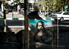 تابلوهای فرش دستباف در خیابان ولی عصر تهران و انعکاس دوچرخه سوار در حال عبور از پیاده رو خیابان.