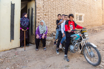چند نوجوان سوار بر موتورسیکلت در کنار دو زن سالمند مقابل یکی از خانه‌های روستای برزنون در فصل تابستان.